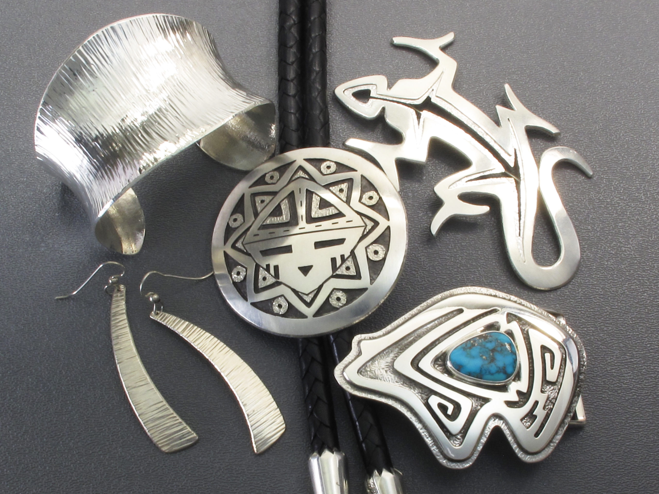 Grouping of sterling silver jewelry on a grey background showing hammer textured cuff and earrings, an overlay medallion bolo tie, pierced silver lizard pin, and an overlay stylized bear buckle set in the center with a turquoise cabochon by Duane Maktima..