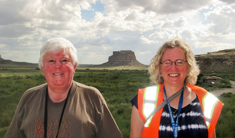 Dennis Gilpin on the left with short white hair and Dr. Terry Hays-Gilpin on the right with shoulder length blonde hair smiling next to each other outside with desert and mesas in the background